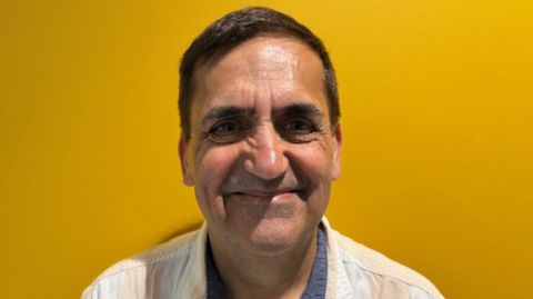 Vineet Vijh is looking at the camera and smiling. He has short, dark hair and us wearing a beige top. Behind him is a mustard-coloured wall.