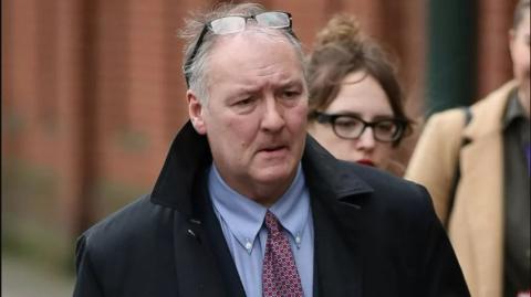 A man standing outside. He has grey hair and glasses perched on his head. He is wearing a black coat, a blue shirt and a maroon and white and blue tie.