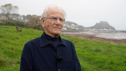 John Henwood - a man wearing rimless rectangle glasses, a navy collared overshirt and a navy turtle neck. He is standing outside on grass with the beach and houses in the background.