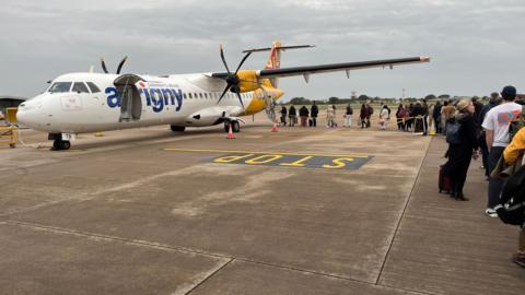 The image shows an Aurigny ATR aircraft with a queue of passengers snaking towards the plane. The aircraft has two propellers. Its tail is painted yellow while the rest of the fusillage is white. The plane has the word Aurigny on the side but the baggage door is open so the U is not visible. 