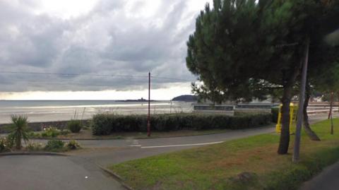 Screenshot taken from Google Maps of La Route De La Haule over looking the beach where the new cafe would be built. The image shows an area of grassland to the right with several trees in a line. The public toilets can be seen behind the trees. The walking and cycling track passes between the trees and the toilets.