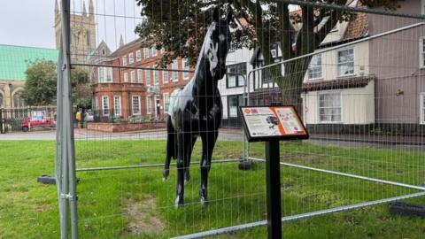 A fibreglass black horse, painted with images of Black Beauty and author Anna Sewell, sited with an information board, outside Great Yarmouth Minster and the house where Ms Sewell was born. The image shows the horse leaning to its left.
