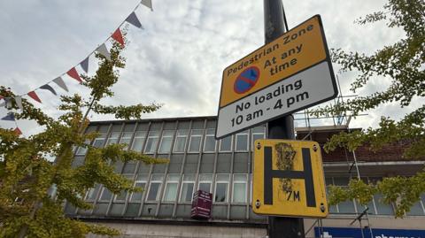 Pedestrian zone warning sign attached to a post in Spalding town centre. There is a mid-20th Century office block and trees in the background.