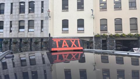 A red taxi sign in top of a black car. The reflection of several tall buildings can be seen in the paintwork.