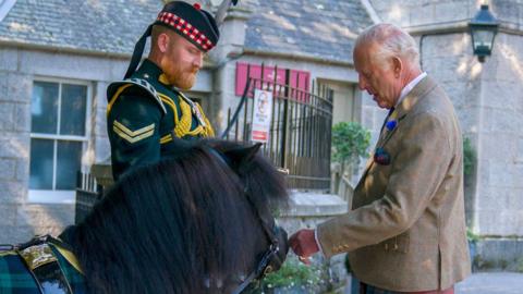King Charles III - who is wearing tweed jacket, matching waistcoat, white shirt and blue tie; kilt, black shocks and black shoes - meets Shetland pony Cpl Cruachan IV (mascot of the Royal Regiment of Scotland) during an inspection of the Balaklava Company, 5th Battalion, The Royal Regiment of Scotland, at the gates of Balmoral, as he takes up summer residence at the castle.