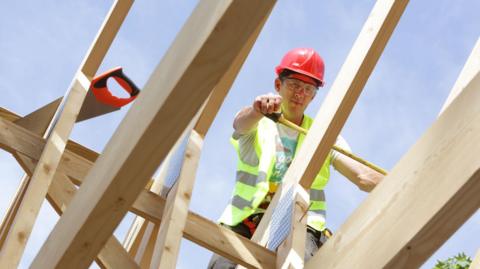 A man in a yellow hi-vis jacket and red hard hat is on top of a wooden house frame, using a measuring tape. A saw is balanced on the beams next to him and he has protective goggles on 