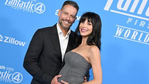 Gruffudd standing with Wallace at The Fantastic Four: First Steps World Premiere. Gruffudd is standing on the left, wearing a black suit. He has one hand on Wallace's bump and the other hand is behind her back. Wallace wears a bandeau grey dress and has black straight hair. 