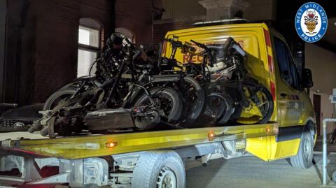A yellow recovery type truck with a long flat bed. On it are at least six e-bikes and or scooters. it is night time so surroundings are dark