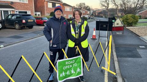 Scott Brooks, is wearing a blue coat with his initials on the bottom of it, with black tracksuit bottoms and a blue and red hat. Next to him on the right is Clare Wallace, wearing a hi-vis vest, green and blue scarf and a black coat. They're stood behind a black and yellow barrier with a green sign on it which reads 'road open to' followed by pictures of people walking, on scooters, on mobility scooters and bikes. Behind them is a residential street and empty road.