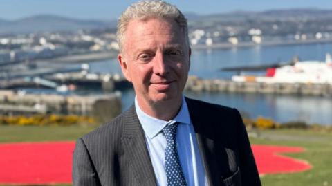 Alex Allinson stood outside in the Isle of Man, a town is visible in the background. He is smiling. He is wearing a pinstripe suit, blue shirt and has short silver hair.