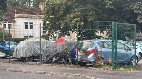 A number of cars in a garage yard. Two vehicles are covered in tarpaulin and a third - a small blue Vauxhall - has scorch marks on its rear. Houses and trees can be seen in the background.