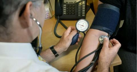 A GP has a stethoscope in his ears as he takes a patient's blood pressure. He has a white shirt on. Opposite him the patient is wearing the blue testing sleeve on their upper arm, and the GP is looking at a pressure gauge. A telephone can be seen on the desk they are sitting at.