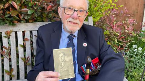 Alan Kennet holding up a picture of himself during 1942 in the RAF. He is wearing glasses and has a white beard.