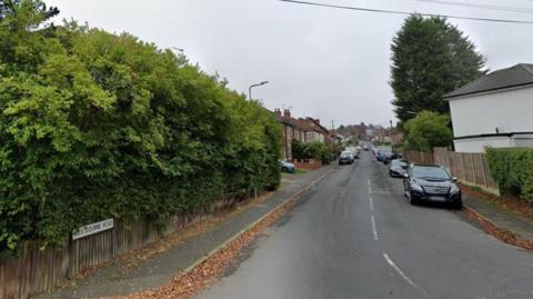 Westbourne Road in Woverhampton with shrubs, a wooden fence and a road sign at the junction and then rows of houses and parked car further up the street.