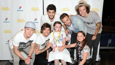 Zayn Malik, Niall Horan, Louis Tomlinson, Liam Payne and Harry Styles of One Direction pose for photographs with Shannon O'Reilly at Wembley Arena. They are posing against a backdrop that says Rays of Sunshine in yellow letters