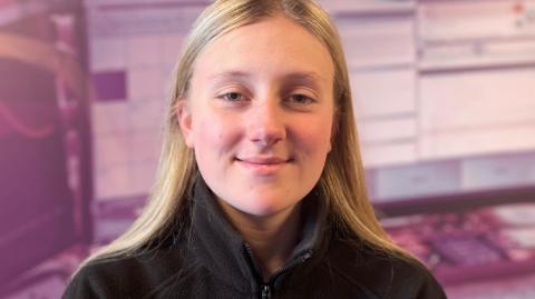 Gabriella Corkish, a young woman with blonde hair neatly behind her ears, she smiles and wears a blakc quarter zip, there is a purple background. 