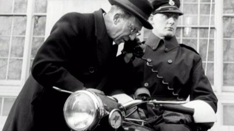 A black and white image of Sir Harold Scott as he leans over a Met Police motorbike to use the receiver of the new two way receiver.  The rider is astride the motorcycle.