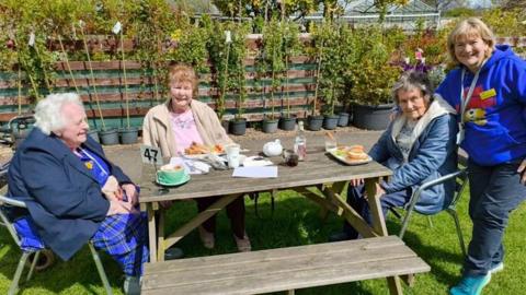 Three elderly women sit around a picnic bench on a lawn at a garden centre surrounded by trees for sale. A carer has her arm around one of the women and is wearing a blue Paddington Bear hoodie.