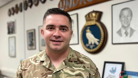 Craig is in camouflage military uniform stands in an office-like room with framed photographs, plaques, and a large wooden sign reading 'UNDAUNTED' featuring a bird crest. A table with various objects, possibly models or decorations, is visible in the foreground.