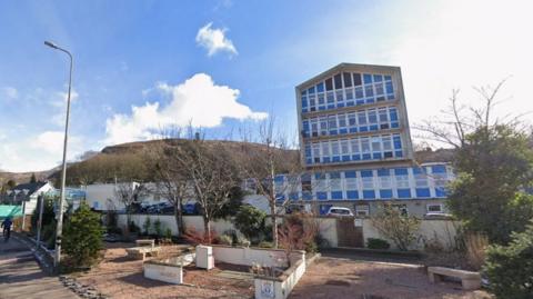 Picture of the outside of Belford Hospital, a multi-storey building painted blue and white. There is a hill in the background, and it is a sunny day with some clouds in the sky.