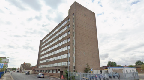 A large, multi-storey office building. Cars are parked on the street outside the building. The sky is cloudy.