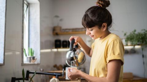 A woman in a yellow tshirt stands at a kitchen sink, pouring water from a silver stovetop kettle into a mug.