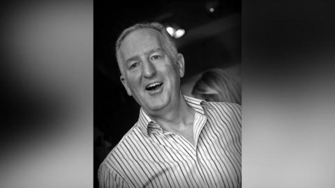 Black and white image of a man smiling with mouth open wearing an open neck white shirt with horizontal stripes of different shades, with the back of a ladies head in the background.