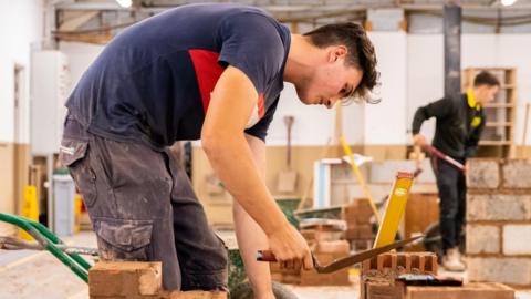 A man holding a trowel is constructing a wall from bricks. He is wearing a red and navy blue top and black cargo trousers. There is another person in the background wearing a black and yellow top and black trousers.