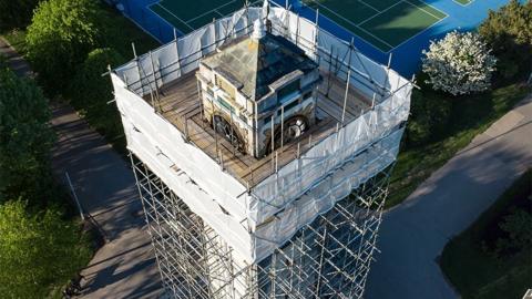 Drone view of the Cocker Memorial Clock Tower covered in scaffolding, with white sheeting to the very top. In the background are gardens, paths and tennis courts