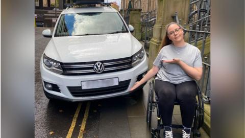 Jennie, a woman in a wheelchair, gesturing towards a white Volkswagen family car which has been parked on a pavement, blocking her from being able to get past without travelling in the road itself.