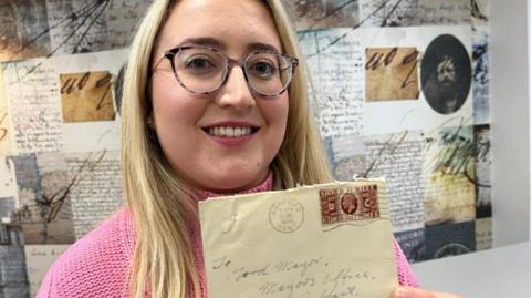 A woman with a long blonde hair wearing a pink jumper and glasses smiling at the camera as she holds an old opened envelope. She is standing in front of a display that shows old documents and photos.