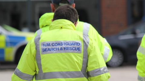 The back of a member of HM Coastguard, they are wearing a bright yellow jacket with the words HM Coastguard Search and Rescue printed on it 