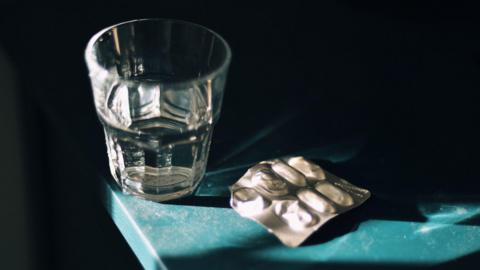 A half-empty medication blister pack next to a glass of water on a dusty table, in a dark room.