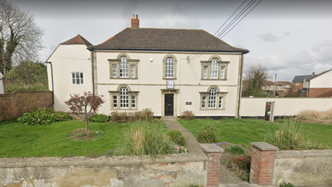 A street view image of a grand-looking beige building with grass and trees in front of it.