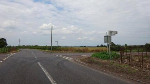 Four roads leading into a crossroads junction. There are green fields on either side of each road. The sky is cloudy, and there is a direction sign in the foreground. 