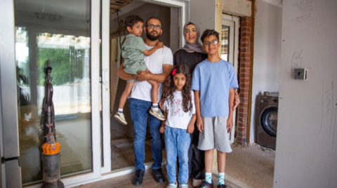 A family of five stand in a doorway. Ahmed Ali has a beard and glasses and is carrying a young boy. His wife Rifat stands next to him and is wearing a head scarf. In front of her is a boy with glasses and a blue T-shirt and a younger girl with long, curly hair and wearing a white T-shirt.