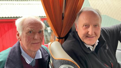 Rod Dolby, a man with white hair, is wearing a green coat with a black collar. He is sitting behind John Bradshaw on a coach. John has grey hair and is wearing a black zipped-jumper. They are both smiling. 