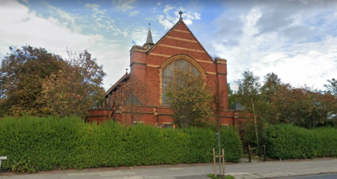 Image shows the red brick St Andrews church in Bootle. It stands behind a large hedge on a main road. 