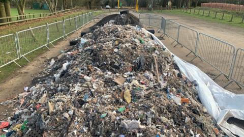 A huge pile of waste dumped in a car park at a park. It has been closed off by metal fencing. The car park has a mud surface and is surrounded by wooden posts and grass.