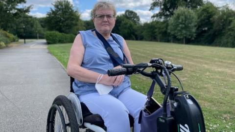 Disability activist Flick Williams pictured in a park, wearing a blue sleeveless top and blue trousers. She sits in a wheeled motorised vehicle. 