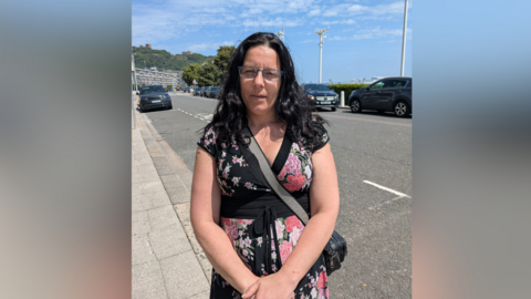 Sarah, from Dover, who took part in the personalised budget trial. Sarah is wearing a cross-body bag and a black/pink dress. She has long black hair and is standing on the pavement. 