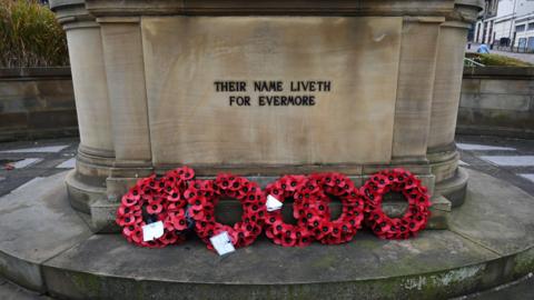 A large stone memorial with several poppy wreaths propped up in front of it. Written in black capital letters on the front of the memorial it says "Their name liveth for evermore".