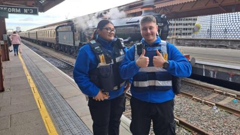 A woman with a long dark ponytail and glasses, and a man with short brown hair and a moustache. They are both in light blue uniform fleeces, standing on a station platform with a steam train behind them. 