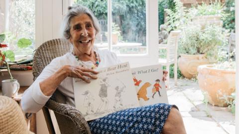 Helen Oxenbury sits in a chair in a conservatory, she is holding two books with a picture of a bear on the cover.