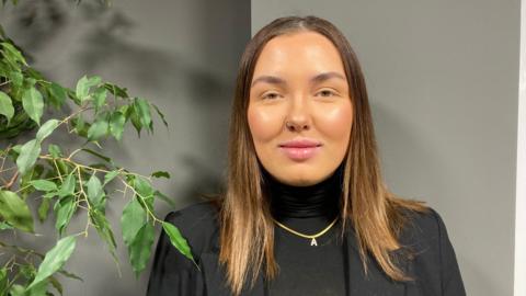 Chelsea Hutton, wearing a black top and blazer. She is wearing a gold necklace with the letter A. She has short brown hair. She is stood in front of a grey wall. 