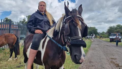 Woman with long blonde hair, blue jacket and jeans, sitting on a horse. There is another horse in the background, some grass and a track with more horses, a vehicle and a person.