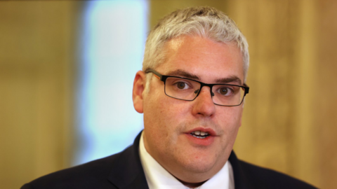 Shows a man with grey hair and black-rimmed glasses in a dark jacket and white shirt.