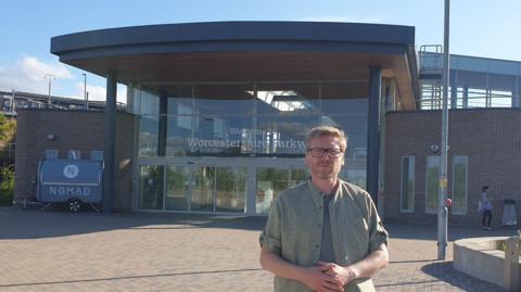 Councillor Dan Boatright-Greene has light brown hair and glasses. He is wearing a grey T-shirt under a green shirt. He is standing outside the front of Worcestershire Parkway, a glass-fronted building with an overhanging roof supported on pillars. 