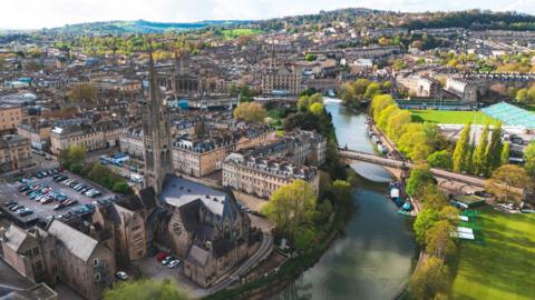 An aerial view of Bath city centre taken by a drone. Landmarks such as the River Avon and Bath Rugby's Rec stadium are visible and it is a clear, sunny day with a range of hills in the background