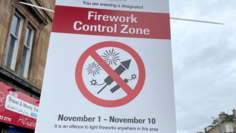 A general view of the Firework Control Zone sign located in the Pollokshields area of Glasgow. It says: "Firework Control Zone: November 1 - November 10. It is an offence to light fireworks in this area." There is a logo featuring two black and white fireworks on a white background with a red circle around it and a red strike through the middle.
